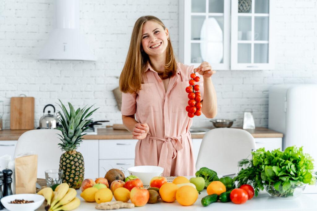 Frau steht in der Küche vor eigens gezüchtetem Gemüse und Obst, hält Tomatenstaude in der Hand.