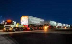 Mehrere, große, beleuchtete Lastwagen, mit Containern beladen, fahren hintereinander bei Nacht. 