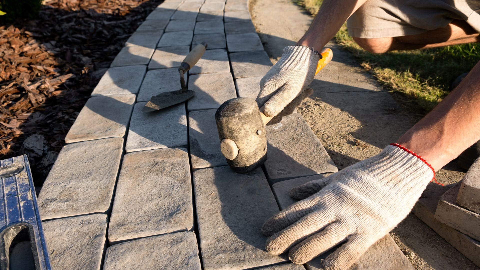 Ein Handwerker kniet am Rand eines neu angelegten Gartenwegs und setzt Kopfsteinpflaster in eine vorbereitete Sandschicht. Neben ihm liegen Gummihammer und Wasserwaage, im Vordergrund eine Schubkarre mit Pflastersteinen. Das Bild zeigt den sorgfältigen Arbeitsprozess beim Anlegen stabiler Wege.